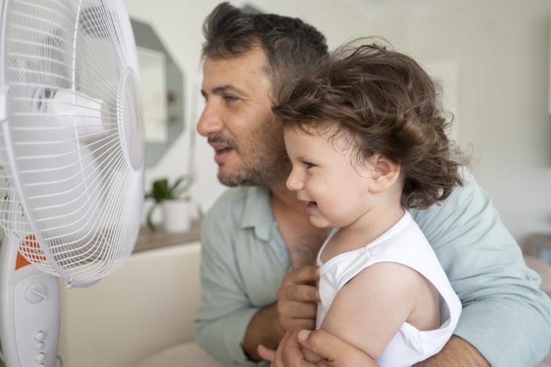 Father and toddler sitting in front of a fan to stay cool.