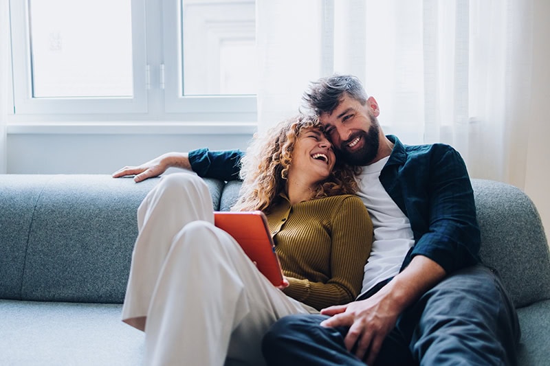 A happy couple sitting close together and laughing on a grey sofa in a brightly lit room.